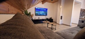 A modern living room with a wooden ceiling, a flat-screen TV mounted on the wall, a sofa, two small round tables, and a plant.