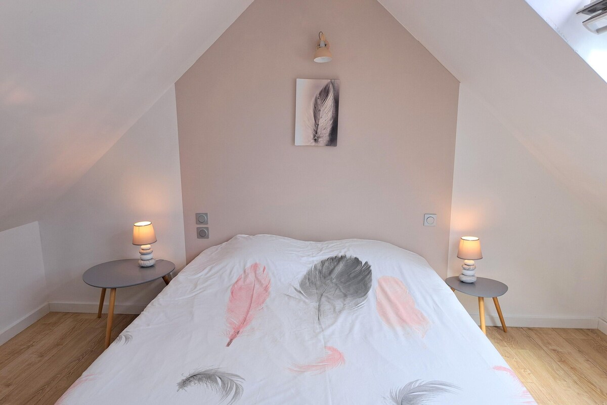 A bedroom with a sloped ceiling, a bed with a feather-themed bedspread, two bedside lamps, and a small painting on the wall.