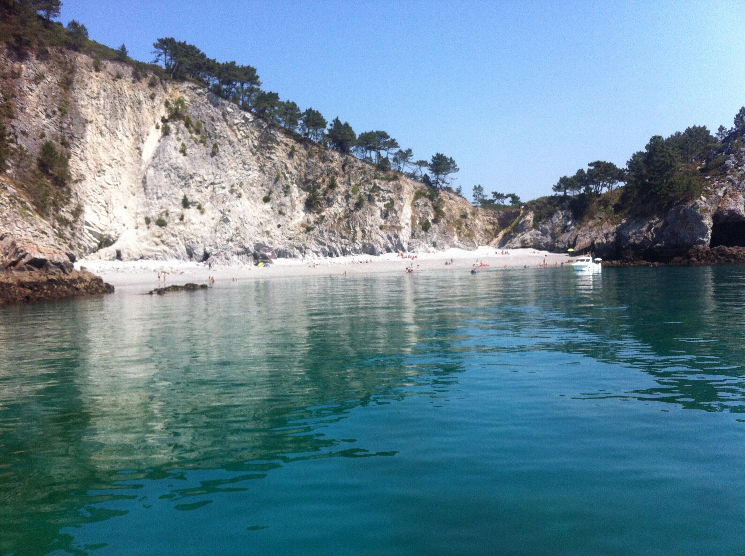 A coastal beach with clear turquoise water, rocky cliffs, and sparse vegetation.