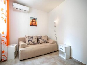 Ein schlicht eingerichtetes Wohnzimmer mit einem beigen Sofa, zwei Beistelltischen, einer Klimaanlage und einer wandmontierten Steckdose.