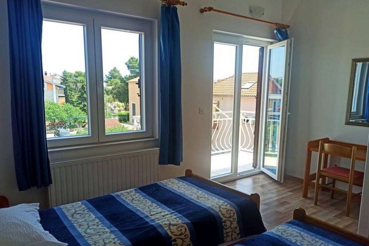 Ein Schlafzimmer mit zwei Betten, einem Fenster mit Vorhängen, einem Sessel und einem Balkon.