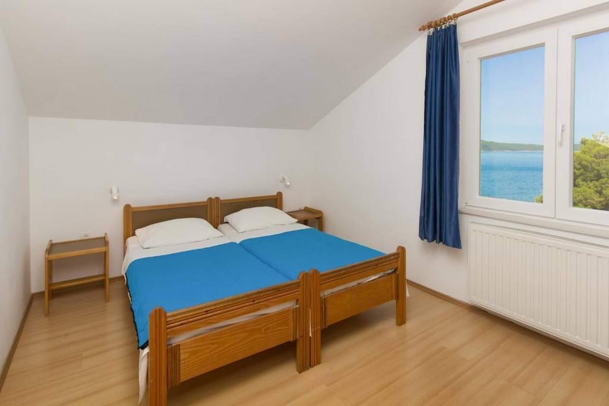 Ein Schlafzimmer mit einem hölzernen Bett, einem Nachttisch, einem Fenster mit Meerblick und einem blauen Vorhang.