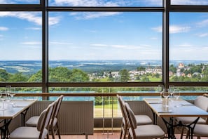 A dining area with a table set for four, large windows offering a scenic view of a town and countryside. - Apartment mit Balkon im Ferienpark in Schoneck (Schoeneck)