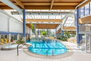 Una piscina interior con techo de madera, paneles de vidrio transparente y sillas de descanso amarillas.