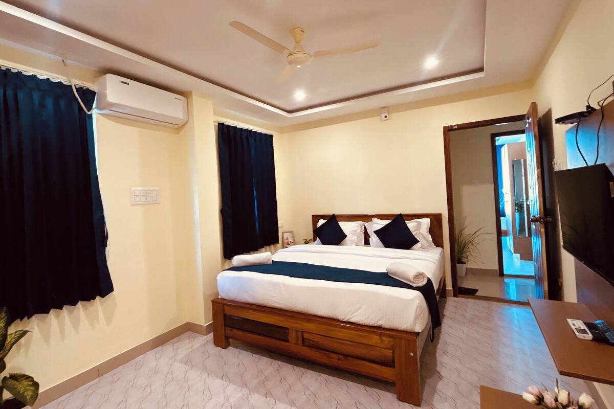 A bedroom with a bed, a ceiling fan, an air conditioning unit, a desk, and a television.