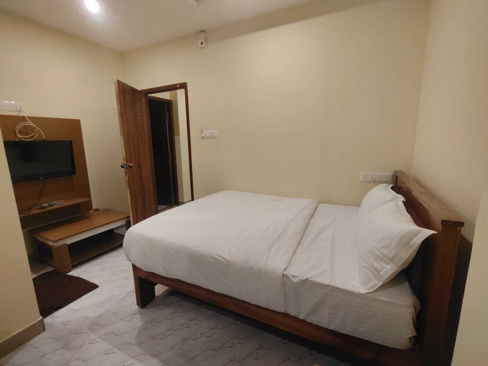 A bedroom with a bed, a TV mounted on a wooden stand, and a door leading to another room.