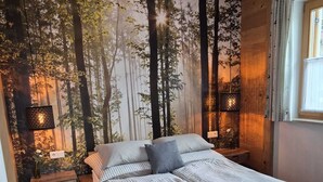 Un lit recouvert d’une couverture à carreaux, un banc, une table de chevet avec une lampe et une fresque murale représentant une forêt.