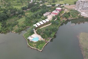 An aerial view of a lakeside property with a swimming pool, a red building, and a large complex in the background. - Lake Alpi Resort Kumbhalgarh (Kumbhalgarh)