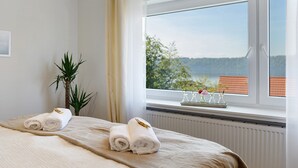 Ein Hotelzimmer mit einem Bett, einer Topfpflanze, einem Fenster mit Blick auf Grünflächen und Gebäude sowie einem Tablett mit Gläsern und Blumen auf der Fensterbank.