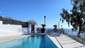 Piscina com água azul cristalina, rodeada por um muro branco e um corrimão. Existem cadeiras de descanso ao longo do corrimão.