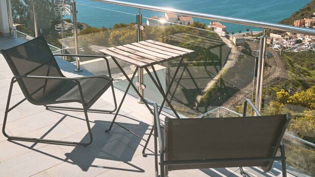 Varanda com duas cadeiras e uma mesa, com vista para a costa, edifícios e uma estrada.