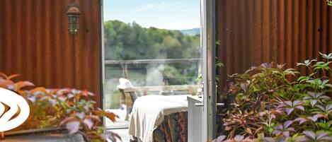 An open door to a balcony with a bed, a table, and a view of a forested landscape.