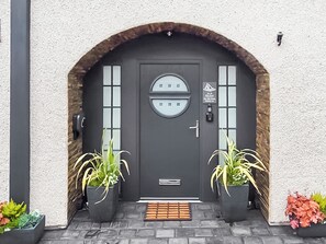 A dark grey door with a circular window, two potted plants, and a small sign above the door.