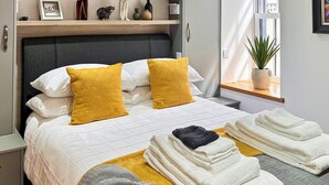 Una cama bien hecha con cabecera, dos almohadas, una colcha y dos toallas dobladas.