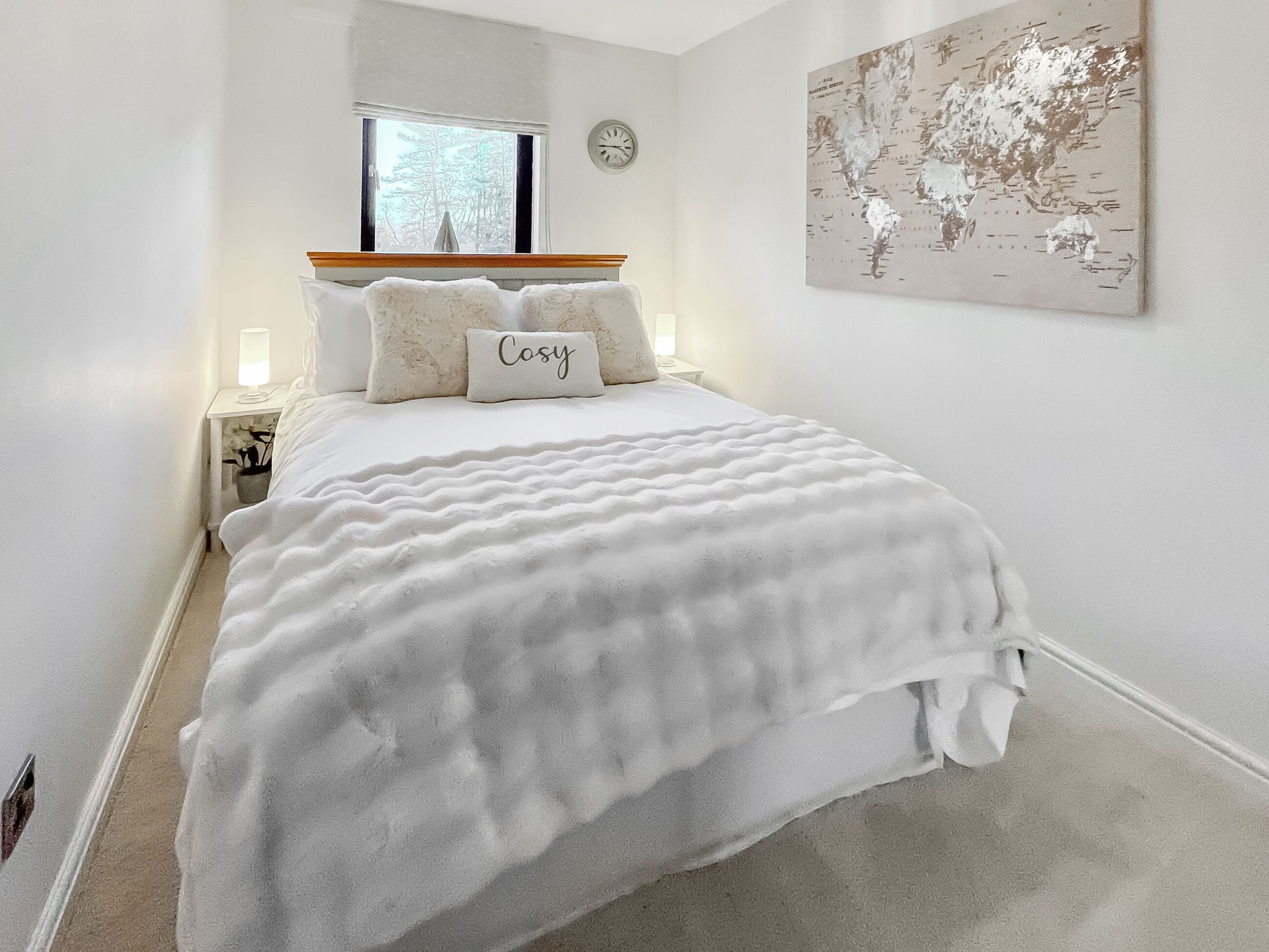 A bedroom with a bed, a nightstand, a lamp, a window with a view of trees, a wall clock, and a world map.