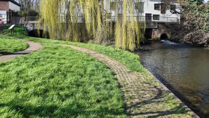 A grassy pathway beside a river with a weeping willow tree, a brick bridge, and a white building with a tiled roof. - 1 bedroom accommodation in Langley (Langley)