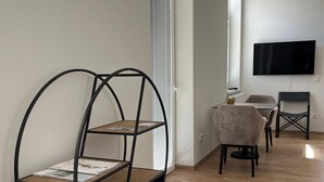 Ett modernt vardagsrum med trägolv, en svart metallstol, ett bord med två stolar, en TV som är monterad på väggen och ett fönster med persienner.