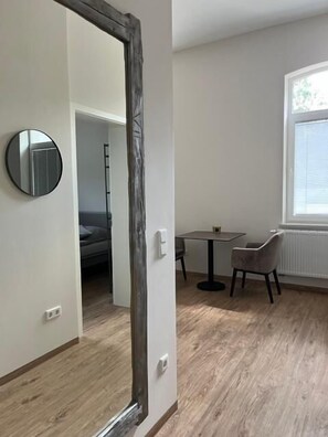 Een kamer met een houten vloer, een ronde spiegel, een tafel met twee stoelen en een raam met jaloezieën.