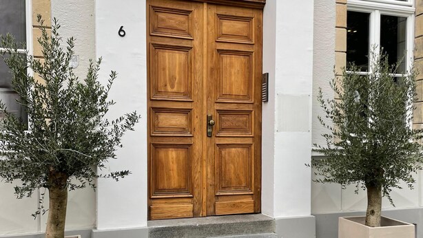 En dubbeldörr av trä med ett glasparti, flankerad av två krukväxter, leder in till en byggnad med en entré som är belagd med kullersten.