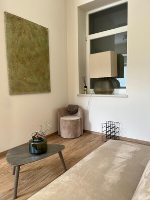 Een minimalistisch ingerichte kamer met een rond poefje, een tafeltje met een vaas, een raam met een lamp en een abstract schilderij dat aan de muur hangt.
