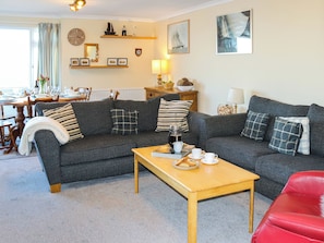 A living room with a sofa, a coffee table, and a red chair. There is a dining table with chairs in the background. - 3 bedroom accommodation in Trearddur bay (Trearddur bay)