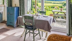 A dining area with a table set for two, a wicker chair, a blue cabinet, and a view of a garden and hills. - 1 bedroom accommodation in Weybourne (Weybourne)