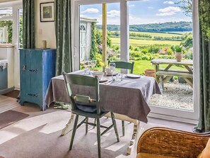 A dining area with a table set for two, a wicker chair, a blue cabinet, and a view of a garden and hills. - 1 bedroom accommodation in Weybourne (Weybourne)