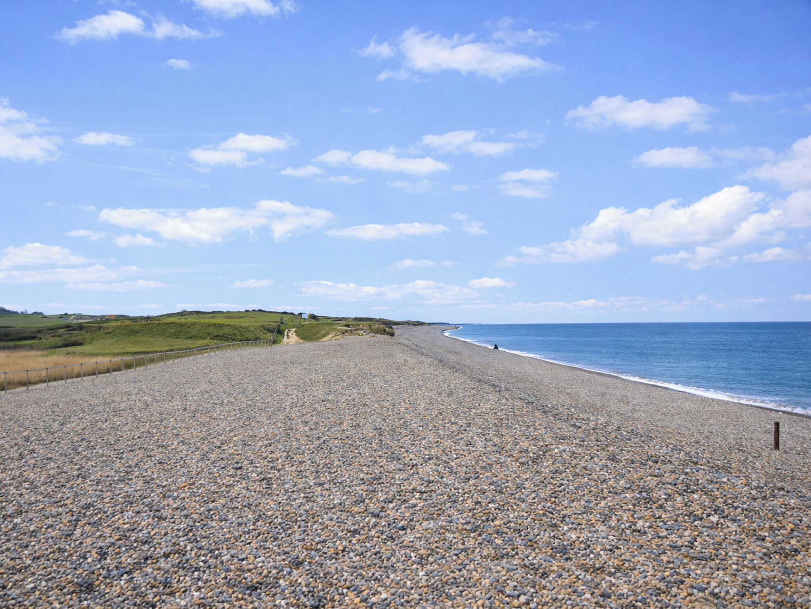 Une plage de galets, avec un ciel dégagé et des collines qui se dessinent à l’horizon.