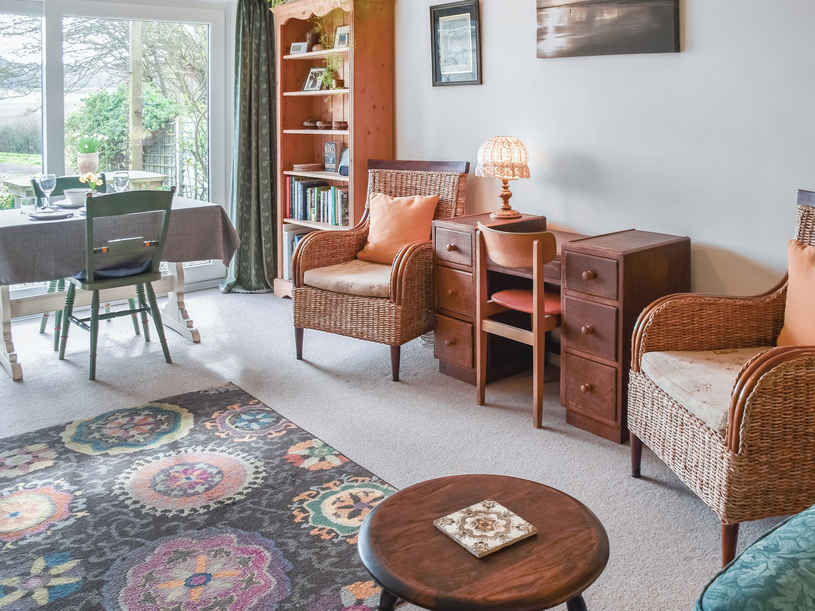 Una sala acogedora con muebles de mimbre, una estantería, una zona de comedor con sillas y una alfombra estampada.