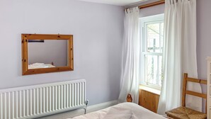 Un dormitorio con una cama, una silla de madera, un ventanal con cortinas, un radiador y un espejo en la pared.