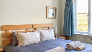 Ein ordentlich bezogenes Bett mit blauer Bettdecke und zwei aufgerollten Handtüchern, ein hölzernes Kopfteil, ein Nachttisch mit Lampe und ein Fenster mit Vorhängen.