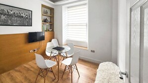 Una habitación moderna con una pared de acento de madera, un televisor de pantalla plana, una mesa de vidrio con sillas blancas, una ventana con persianas y una alfombra blanca mullida.