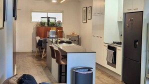 A modern living room with a sofa, a dining area with chairs, a kitchen with a refrigerator, and a ceiling fan.