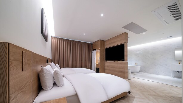 Una camera da letto moderna con una testiera in legno, un letto con biancheria da letto bianca, una TV a parete e un bagno con rivestimento e lavabo in marmo.