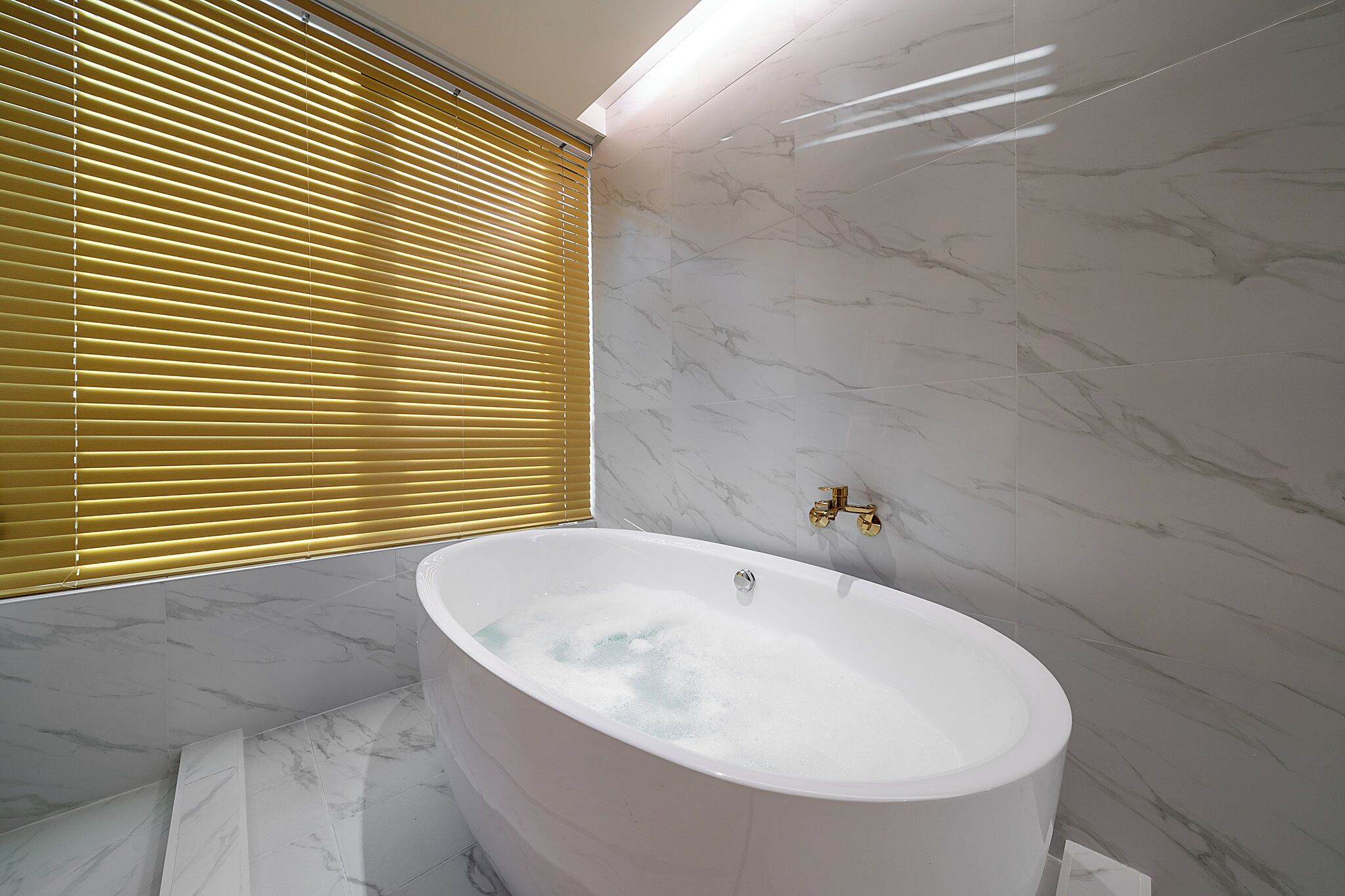A bathroom with a freestanding bathtub, marble walls, and a window with blinds.