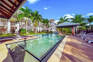 Ein Pool mit Liegestühlen, eine überdachte Sitzgelegenheit und ein mehrgeschossiges Gebäude mit Balkonen.