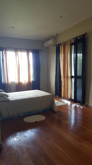 A bedroom with a bed, a window with curtains, an air conditioning unit, and a wooden floor.