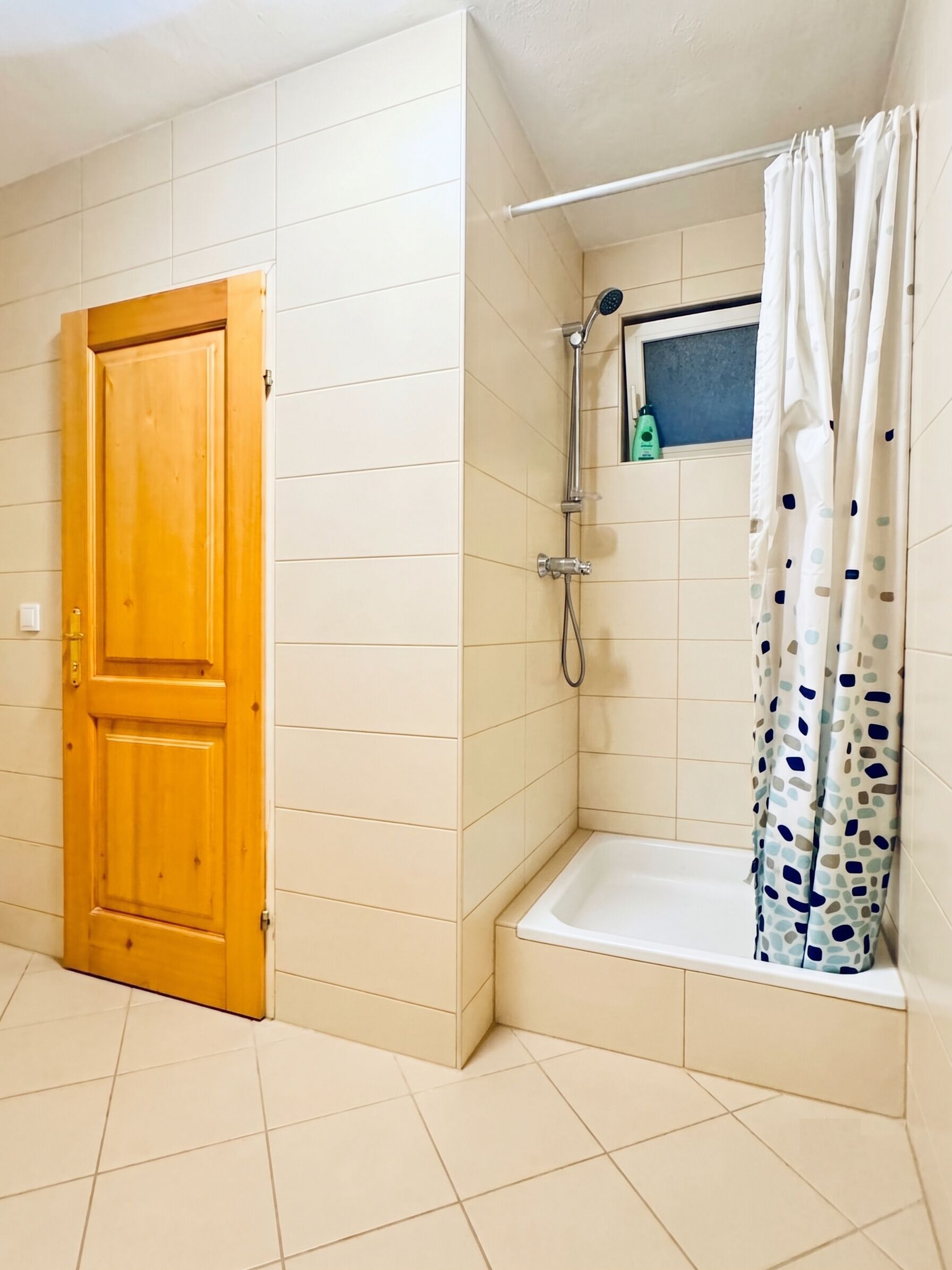 A bathroom with a wooden door, a shower with a curtain, and a small shelf with toiletries.