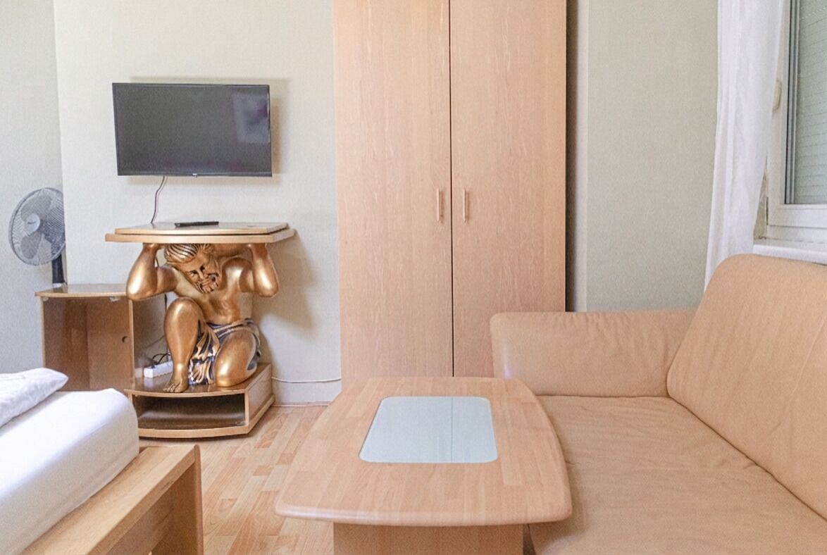 A room with a bed, a sofa, a small table with a flat-screen TV, a wooden cabinet, and a fan.