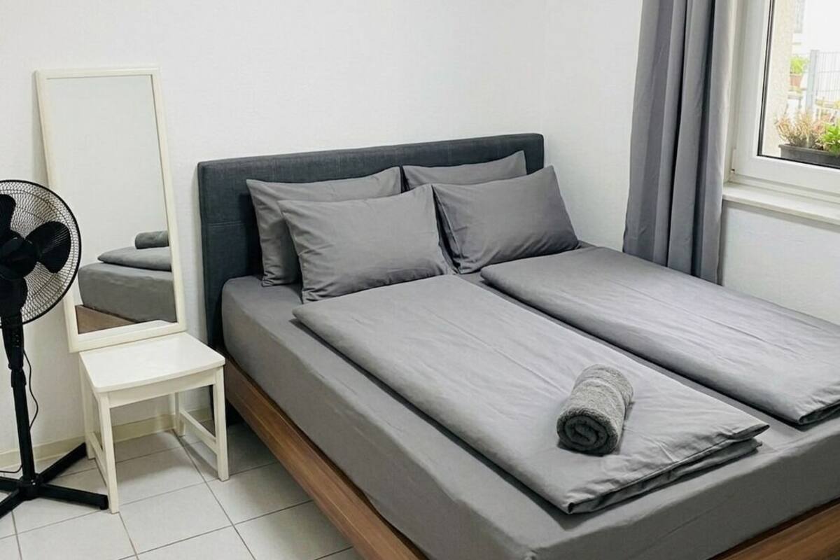 A single bed with grey bedding, a small bedside table, a standing fan, and a window with curtains.