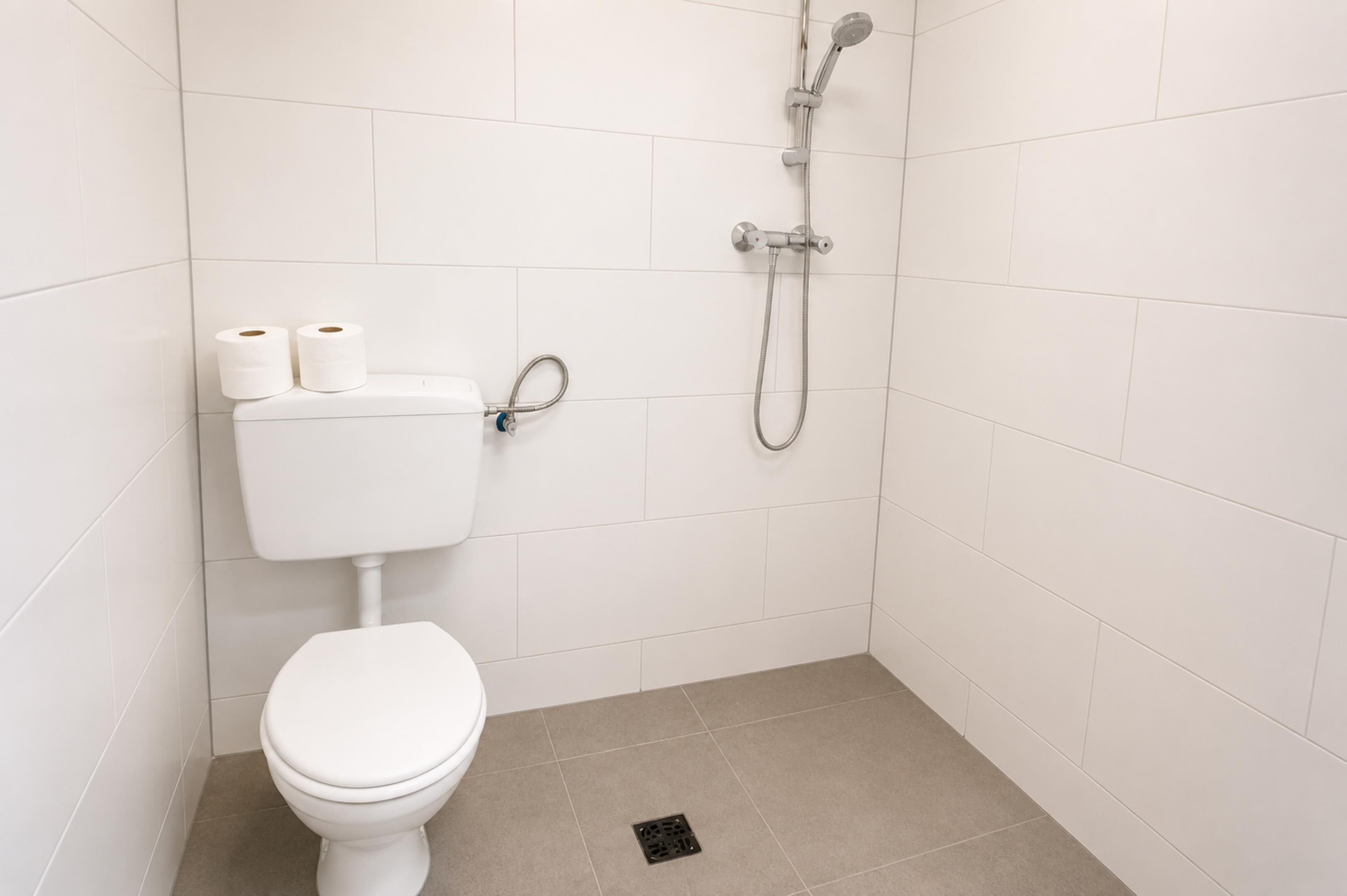 Un baño pequeño y blanco con un inodoro, dos rollos de papel higiénico, un cabezal de ducha y una manguera de ducha.