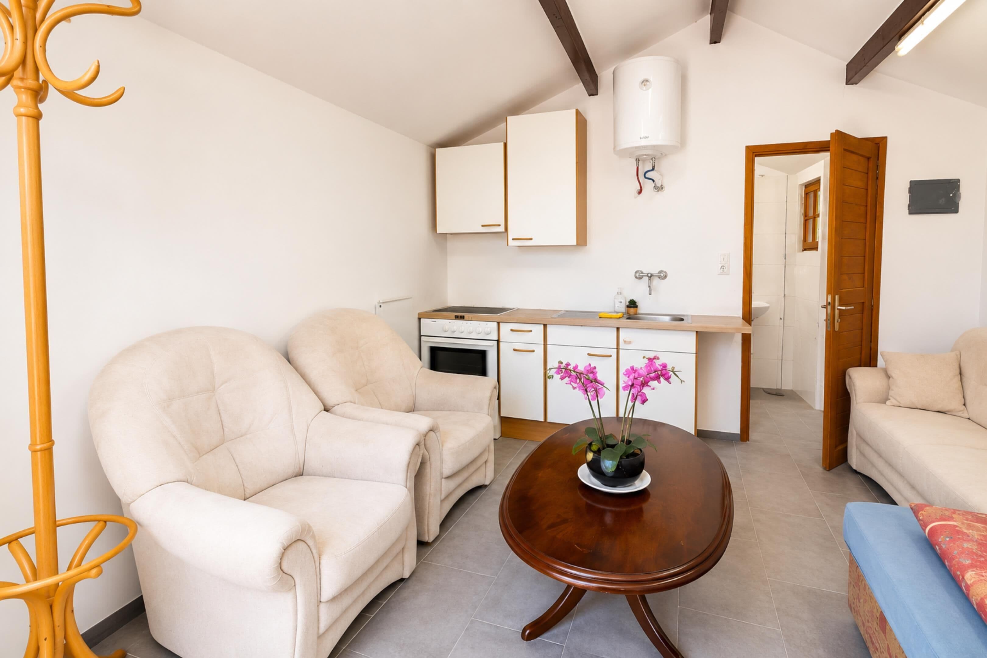 Un espacio habitable compacto con kitchenette, dos sillones color beige, una mesa de centro de madera y una mesita con flores rosadas.
