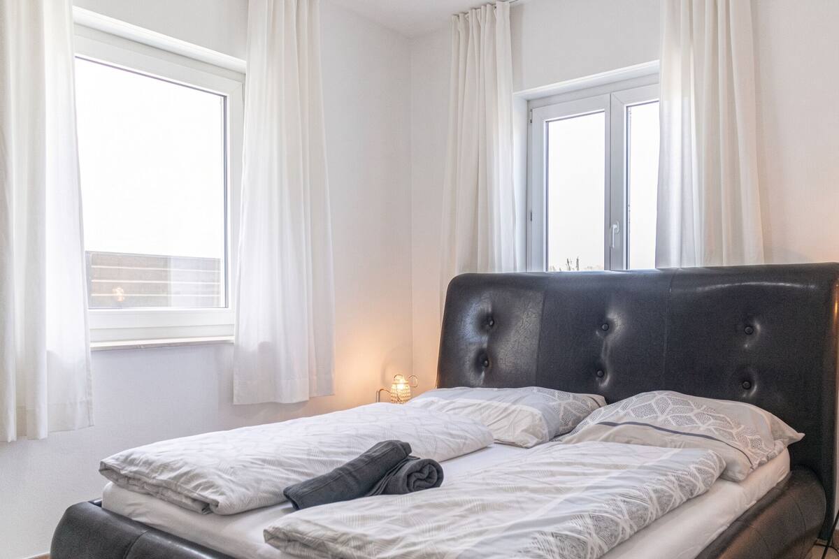 A neatly made bed with a headboard, white curtains, and a small bedside lamp.