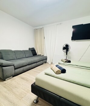 Ein Zimmer mit grauem Sofa, einem Bett mit grünen Laken, einem Standventilator und einem an der Wand befestigten Fernseher.