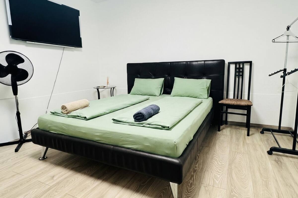 A bedroom with a bed, a standing fan, a TV, and a chair.