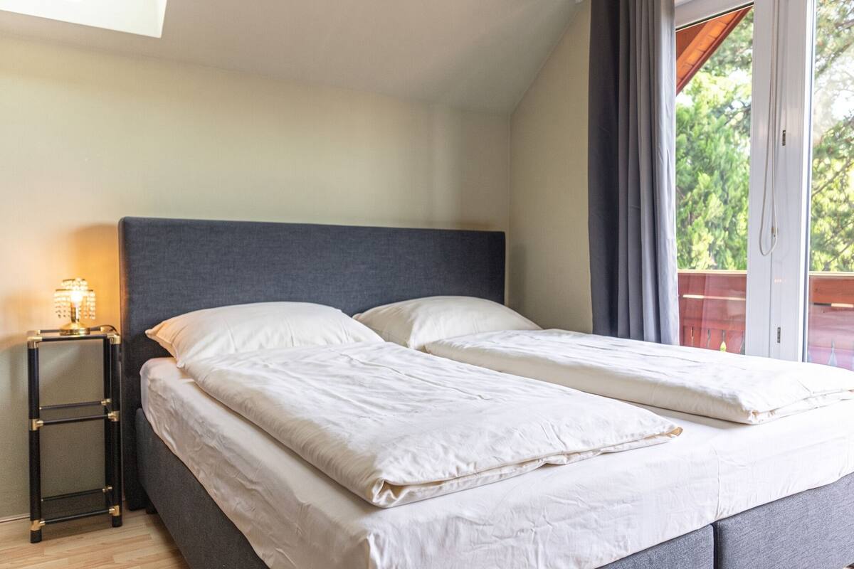 A double bed with a grey headboard, a bedside table with a lamp, and a view of greenery through a window.
