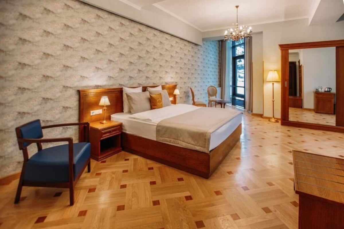 A hotel room with a large bed, wooden headboard, bedside table with lamp, chair, and a mirror.