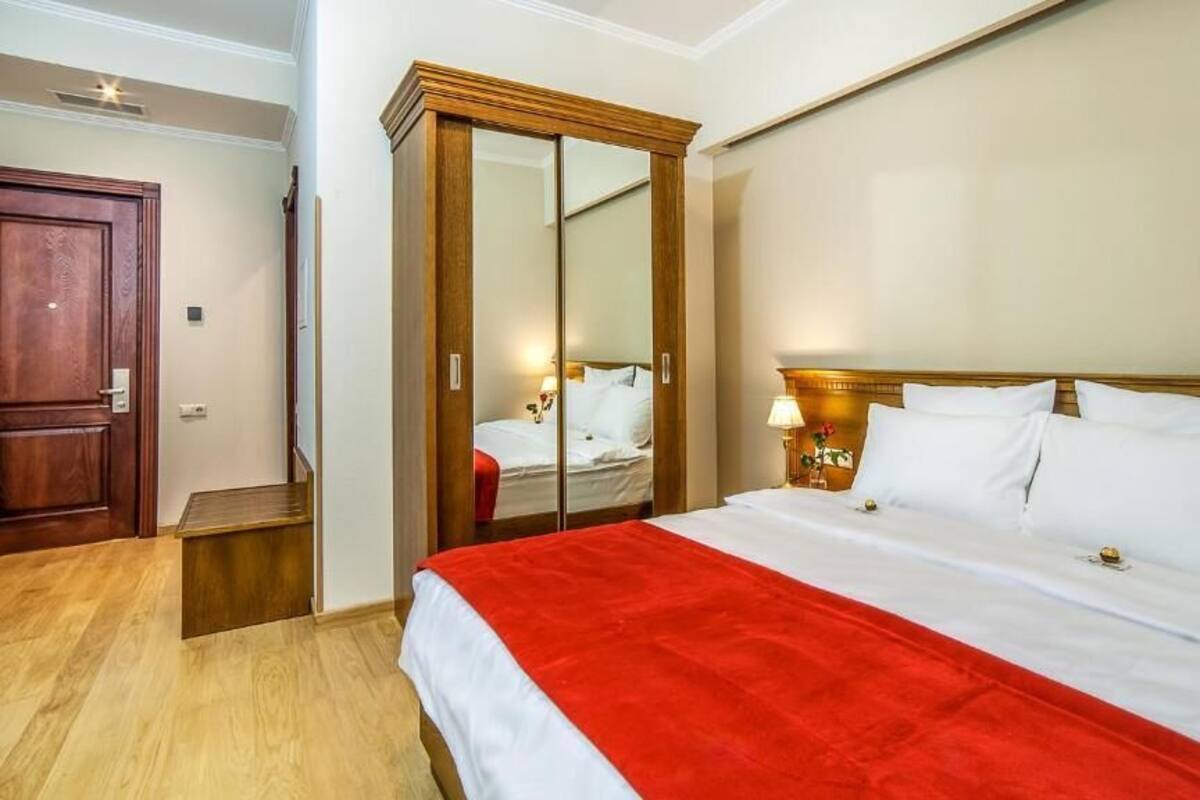 A hotel room with a wooden wardrobe, a bed with white bedding, a red throw, and a bedside lamp.