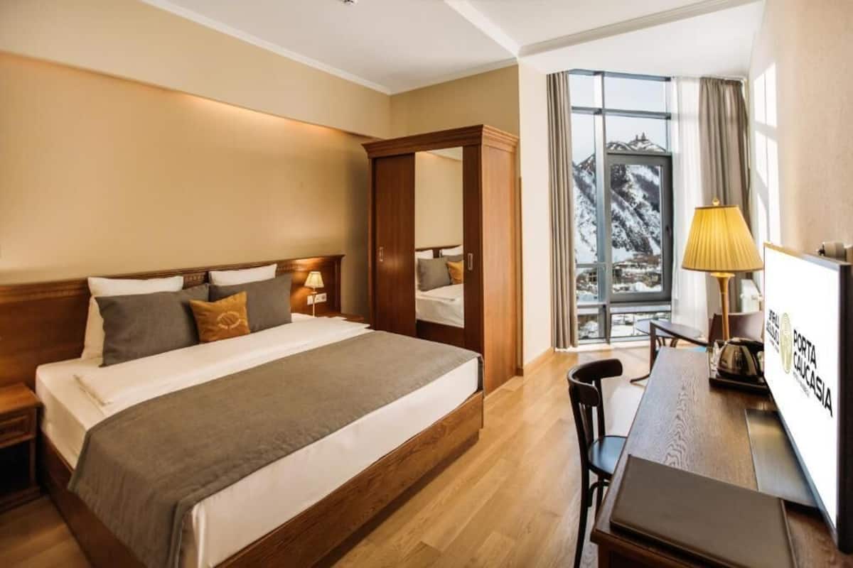 A hotel room with a bed, bedside tables, a desk, and a view of mountains through a window.