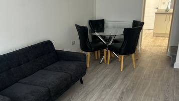 A modern living room with a dark grey sofa, a glass-top dining table with four black chairs, and a wooden floor.